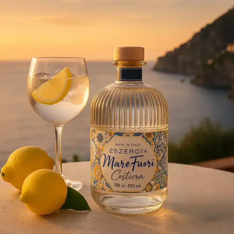 Una bottiglia di vetro chiaro di distillato Mare Fuori Costiera con un'etichetta decorativa e un tappo di legno, accanto a un bicchiere della bevanda guarnito con una fetta di limone e due limoni interi.