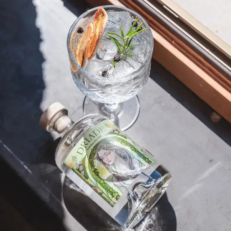Bottiglia di Driade London Dry Gin accanto a un gin tonic con ghiaccio, rosmarino, bacche di ginepro e scorza d’arancia, in luce naturale su un davanzale.