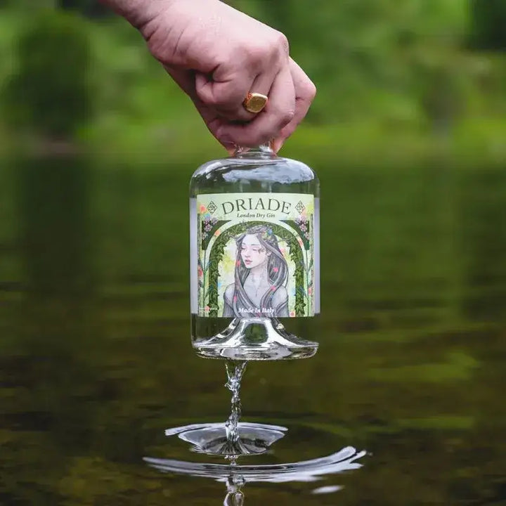 Mano che solleva una bottiglia di Driade London Dry Gin da un ruscello, immersa nella natura, con riflesso sull'acqua e sfondo verde boschivo