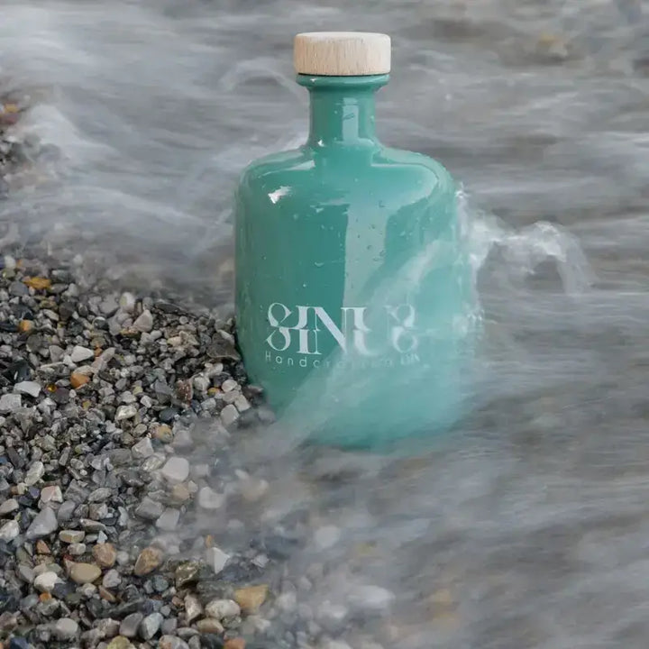Bottiglia di Sinus Gin circondata dalle onde su una spiaggia di ciottoli – un gin che racconta autenticità e paesaggi alpini.