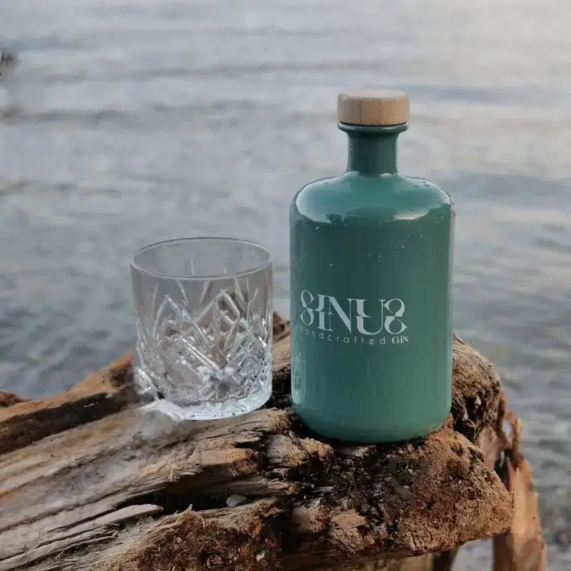 Bottiglia di Sinus Gin artigianale verde smeraldo con bicchiere in cristallo su tronco di legno davanti al lago – un'immagine che esprime natura e artigianalità italiana.